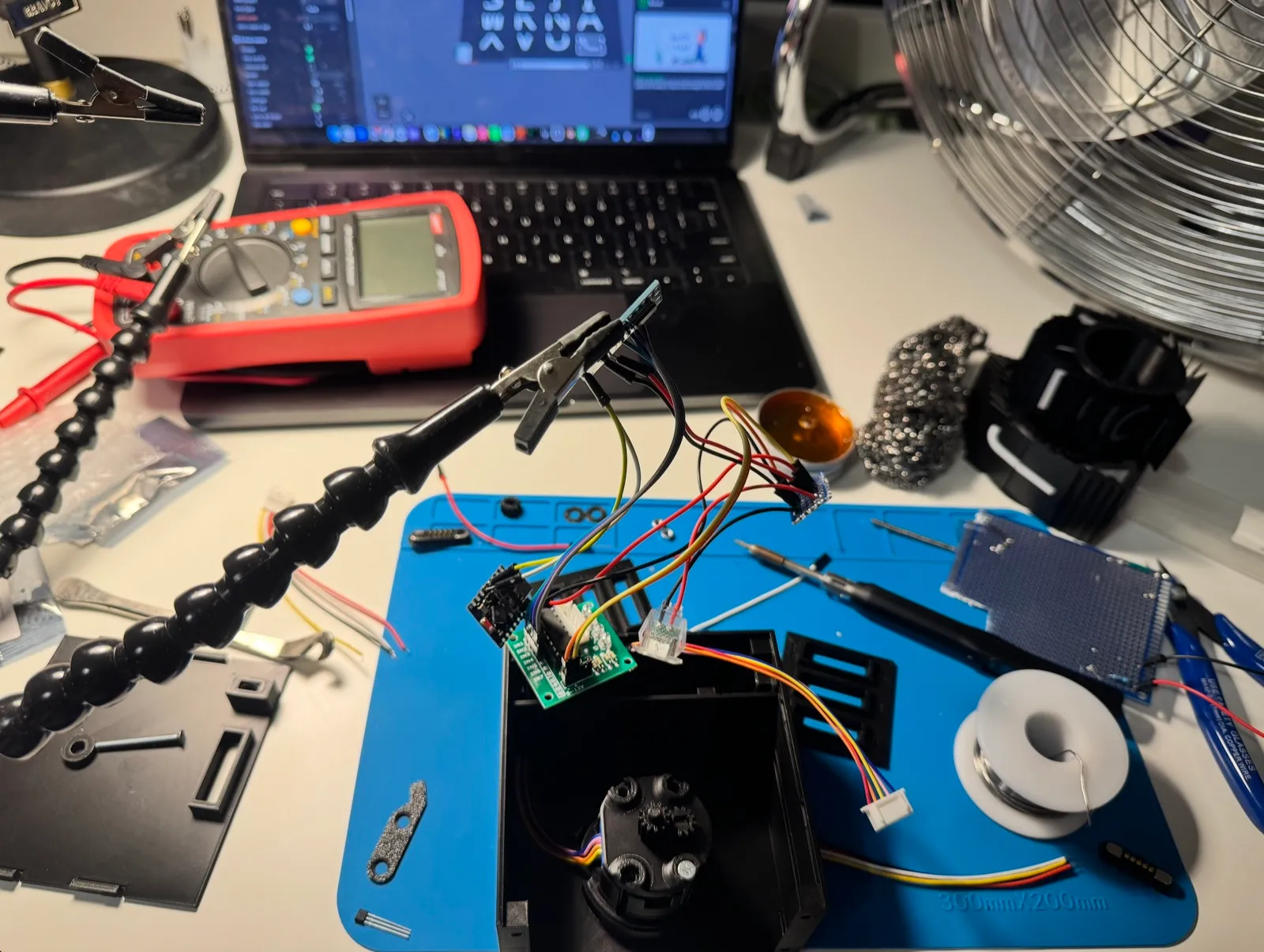 A desk with a blue soldering mat, a small board held in helping hands and wired to a plastic enclosure, a multimeter, a spool of solder and a laptop in the background